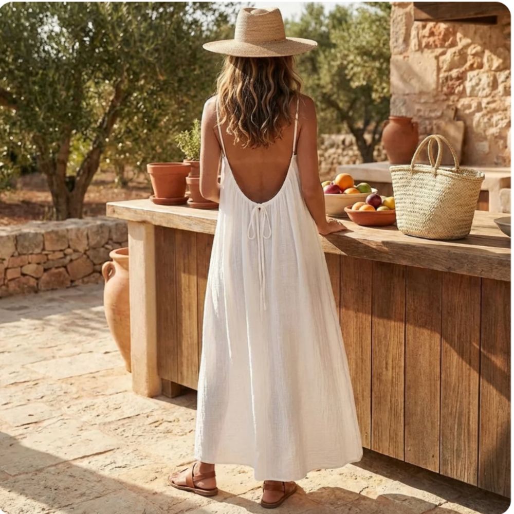 Woman in a ackless_Cami_Maxi_Dress white dress and straw hat standing by a wooden counter outdoors.